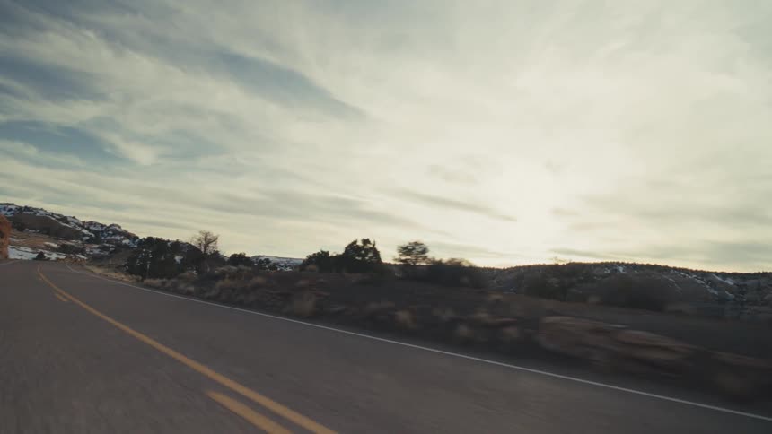 A winding two lane highway through a vast desert valley surrounded by rocky mesas and hills covered in patches of winter snow under a bright overcast sky