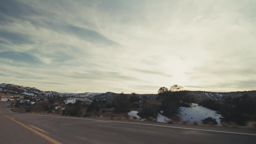A winding two lane highway through a vast desert valley surrounded by rocky mesas and hills covered in patches of winter snow under a bright overcast sky