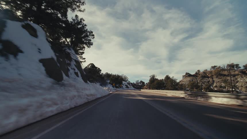 A winding two lane asphalt highway through a mountainous canyon with melting snow on the embankments under a bright blue sky with wispy clouds and scattered evergreen trees