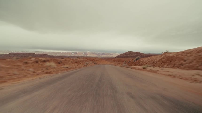 A view from a moving vehicle travels down a straight paved road through an expansive orange desert landscape with rocky cliffs and low plateaus under a heavy grey sky