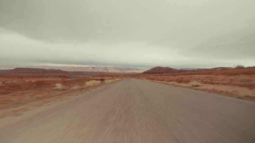 A view from a moving vehicle travels down a straight paved road through an expansive orange desert landscape with rocky cliffs and low plateaus under a heavy grey sky