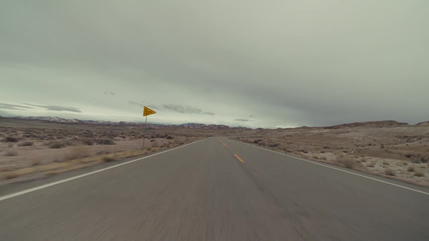 A view from a moving vehicle travels down a long straight road with two yellow lines through a dry desert landscape with scattered bushes and distant mountains under a grey overcast sky