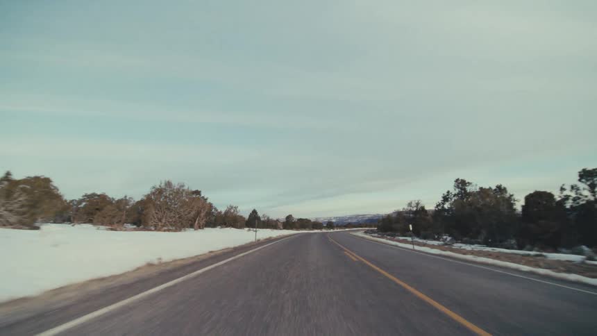 A two lane asphalt road winding through a snowy desert landscape under a pale overcast sky with low shrubs and distant mountains lining the cold winter horizon