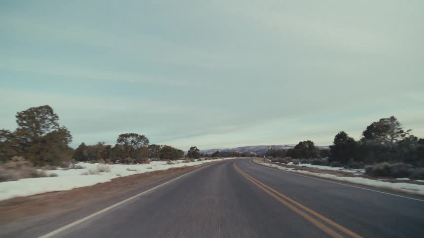A two lane asphalt road winding through a snowy desert landscape under a pale overcast sky with low shrubs and distant mountains lining the cold winter horizon