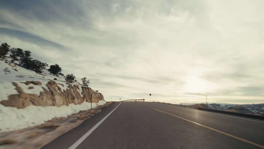 The video shows a firstperson perspective of a vehicle driving along a winding twolane mountain highway the road is surrounded by a rugged desertlike landscape with rocky cliffs and scattered patches of snow under a bright overcast sky