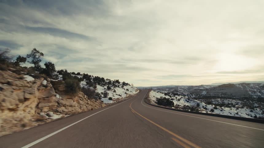 The video shows a firstperson perspective of a vehicle driving along a winding twolane mountain highway the road is surrounded by a rugged desertlike landscape with rocky cliffs and scattered patches of snow under a bright overcast sky