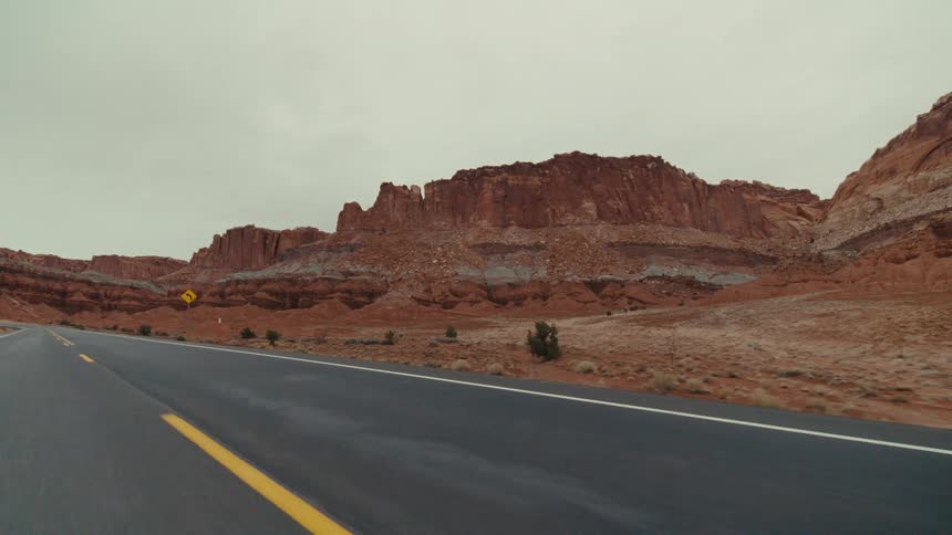 A two lane highway as it curves through a vast desert landscape featuring massive red sandstone cliffs and rocky buttes under an overcast sky with sparse dry vegetation along the side of the dark asphalt road