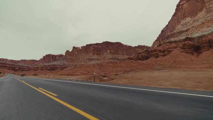 A two lane highway as it curves through a vast desert landscape featuring massive red sandstone cliffs and rocky buttes under an overcast sky with sparse dry vegetation along the side of the dark asphalt road