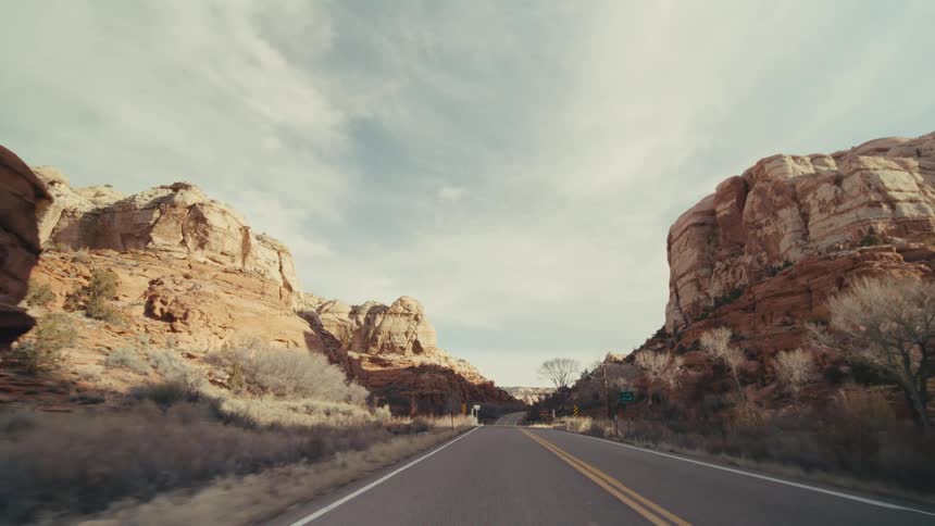 A view from a moving vehicle follows a paved road as it curves past steep orange rock walls and snow covered hills under a bright sky with soft clouds