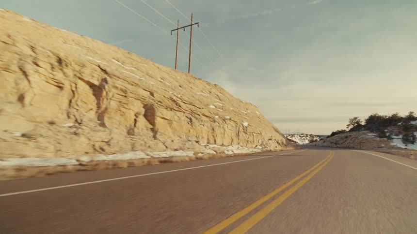 A view from a moving vehicle travels along a curved paved highway with two yellow lines past light brown rocky cliffs and hills with patches of snow under a soft blue sky