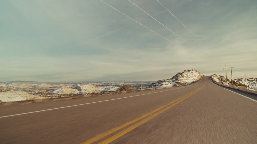 A view from a moving vehicle travels along a curved paved highway with two yellow lines past light brown rocky cliffs and hills with patches of snow under a soft blue sky