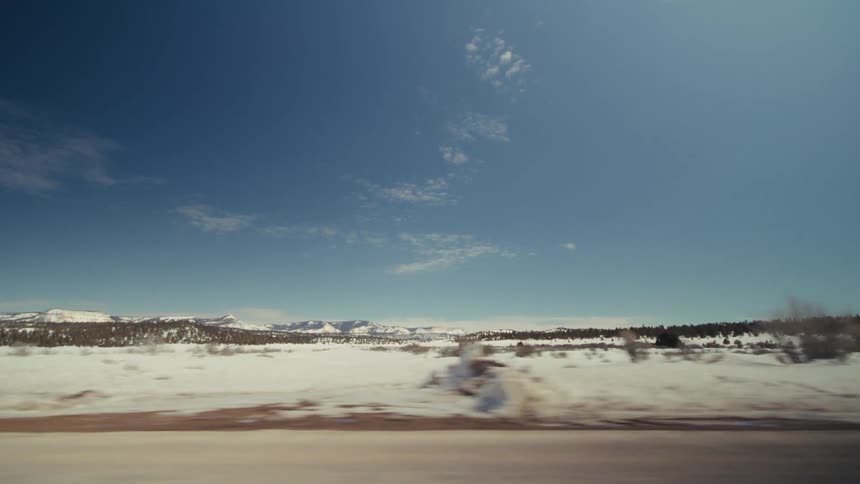 A view from a vehicle moves quickly past a flat snowy field and a line of evergreen trees with distant mountains under a clear blue sky