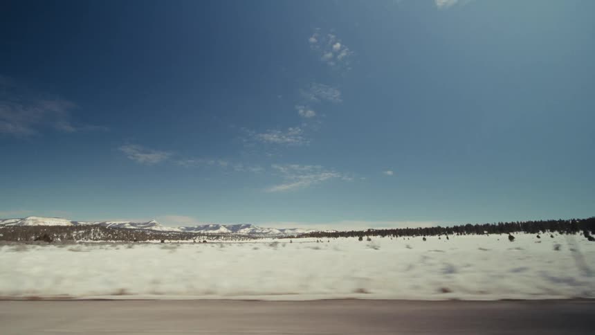 A view from a vehicle moves quickly past a flat snowy field and a line of evergreen trees with distant mountains under a clear blue sky