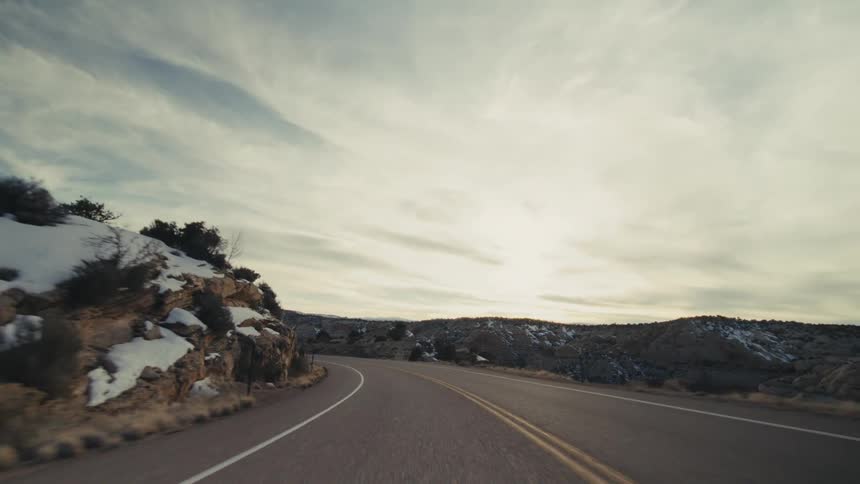 A view from a moving vehicle follows a paved road as it curves past steep orange rock walls and snow covered hills under a bright sky with soft clouds