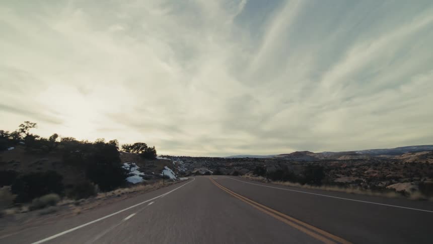 A view from a moving vehicle follows a paved road as it curves past steep orange rock walls and snow covered hills under a bright sky with soft clouds