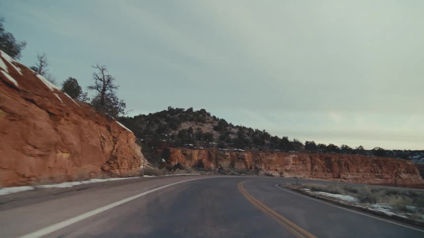 A curved asphalt highway through a winter desert landscape featuring red rock cliffs scattered evergreen trees and patches of white snow under a pale overcast sky