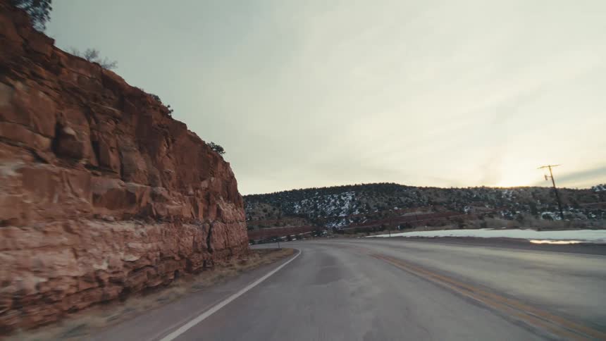 A curved asphalt highway through a winter desert landscape featuring red rock cliffs scattered evergreen trees and patches of white snow under a pale overcast sky