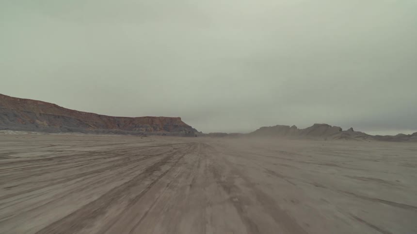 A view from a low camera travels quickly across a vast and flat desert plain driving away from distant brown rocky hills under a dark and heavy overcast sky
