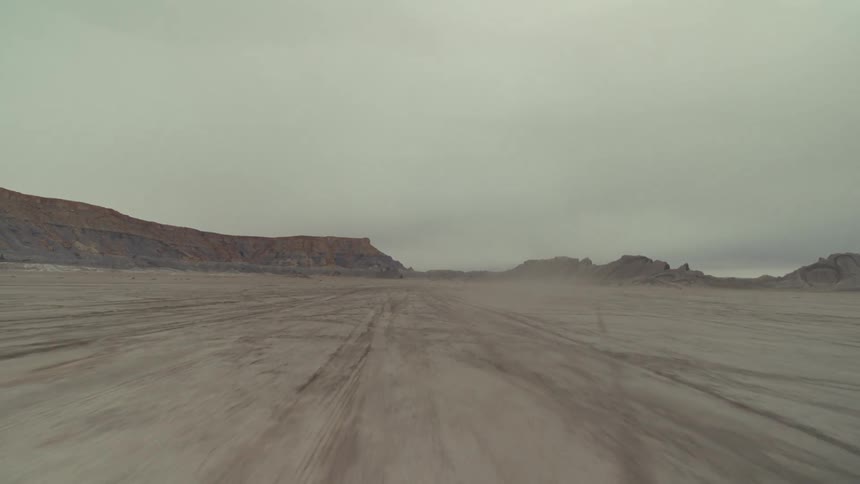 A view from a low camera travels quickly across a vast and flat desert plain driving away from distant brown rocky hills under a dark and heavy overcast sky