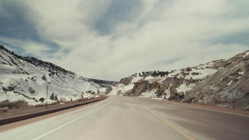 A point of view shot from a vehicle driving on a two lane road through a snowy mountain pass under a cloudy sky