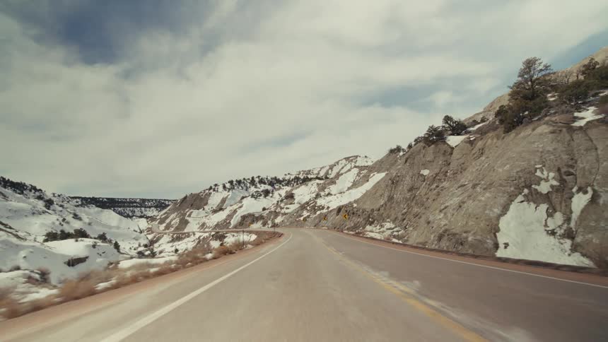 A point of view shot from a vehicle driving on a two lane road through a snowy mountain pass under a cloudy sky