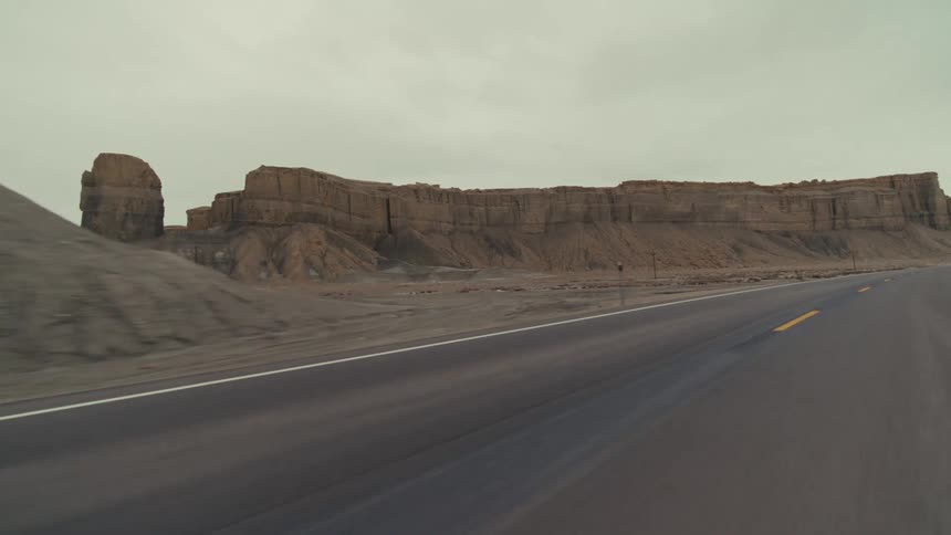 A view from a vehicle moves along a paved highway passing by large rock formations and a desert landscape under an overcast sky
