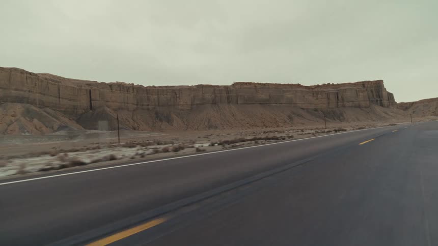 A view from a vehicle moves along a paved highway passing by large rock formations and a desert landscape under an overcast sky