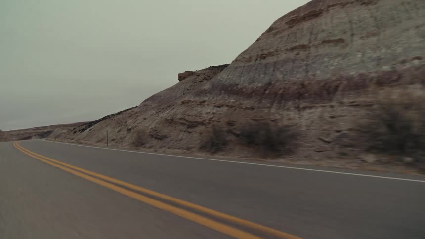 A winding desert highway with double yellow lines passing by steep rocky hills and a metal guardrail under a flat overcast sky