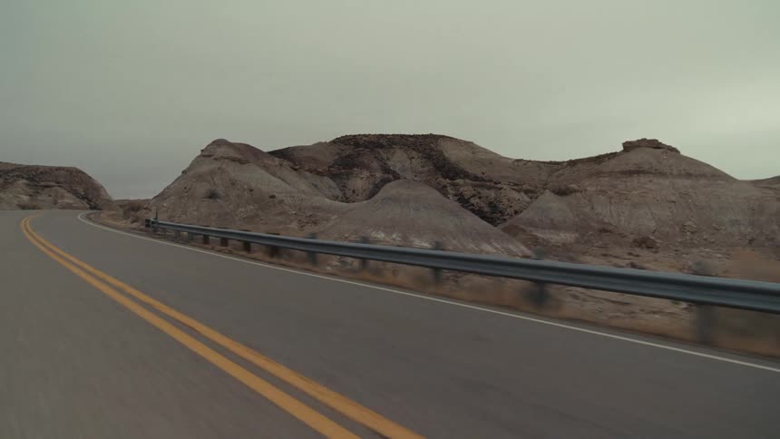 A winding desert highway with double yellow lines passing by steep rocky hills and a metal guardrail under a flat overcast sky