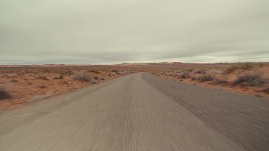 A view from a vehicle travels quickly along a straight paved road through a wide desert landscape with red sand and low bushes under a gray overcast sky