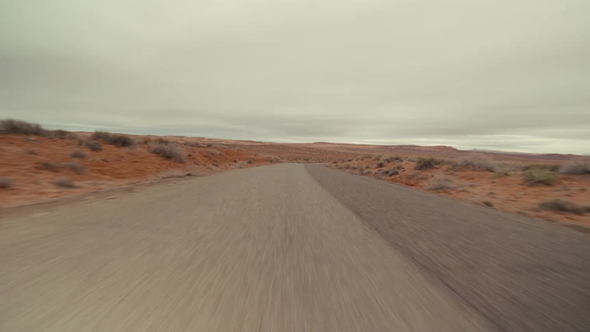 A view from a vehicle travels quickly along a straight paved road through a wide desert landscape with red sand and low bushes under a gray overcast sky