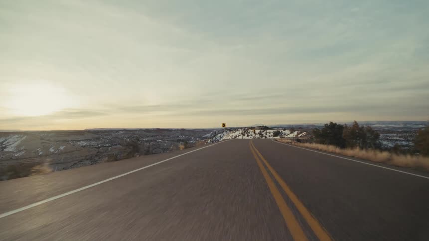 A view from a moving vehicle travels down a winding two lane asphalt road through a hilly desert landscape with patches of snow and scattered trees under a hazy sunset sky