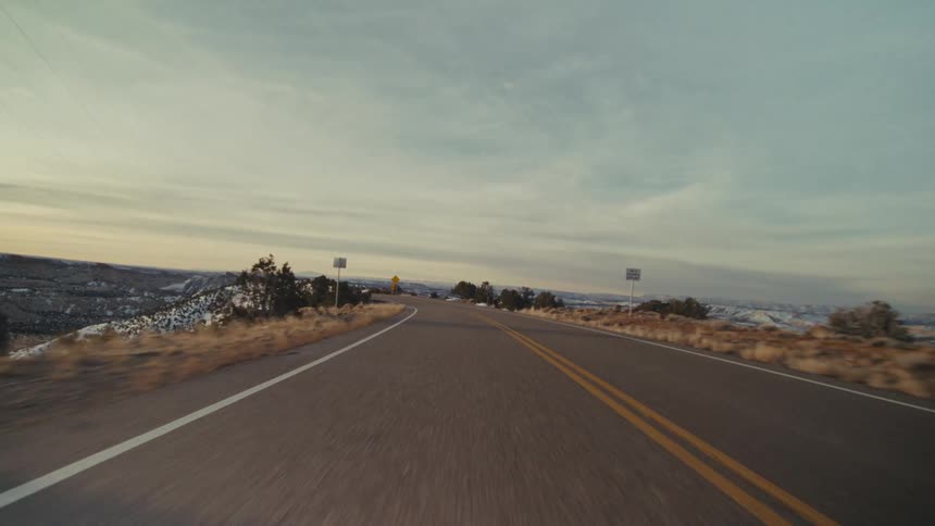 A view from a moving vehicle travels down a winding two lane asphalt road through a hilly desert landscape with patches of snow and scattered trees under a hazy sunset sky