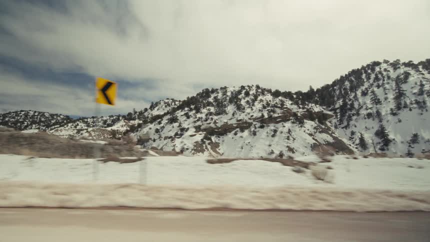 Snowy white mountains with scattered evergreen trees sit under a cloudy sky on a roadside with patches of dry brown grass and dirt in the foreground