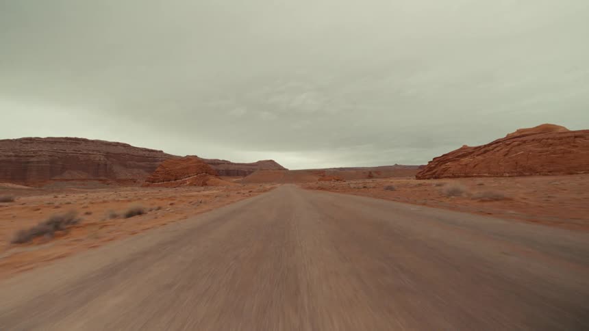 A point of view shot of driving on a dirt road through a desert landscape with red rock formations under a cloudy sky
