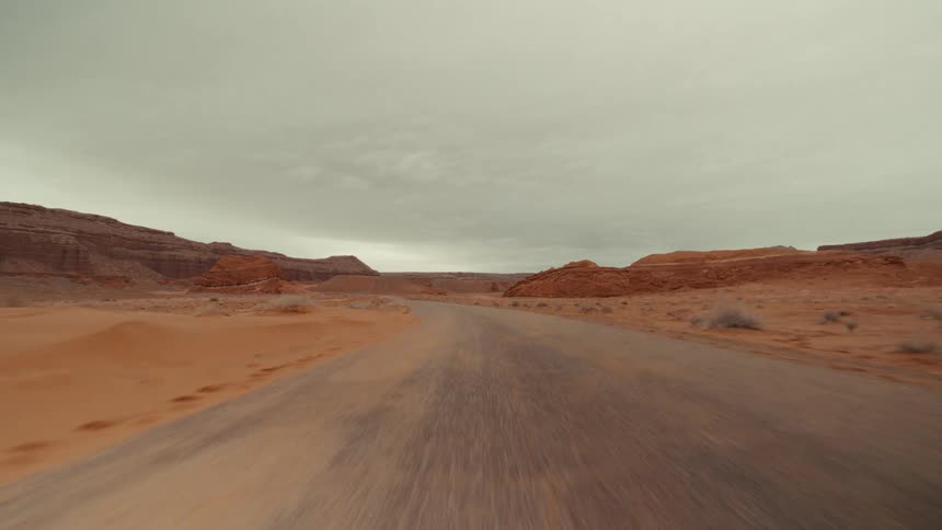A point of view shot of driving on a dirt road through a desert landscape with red rock formations under a cloudy sky