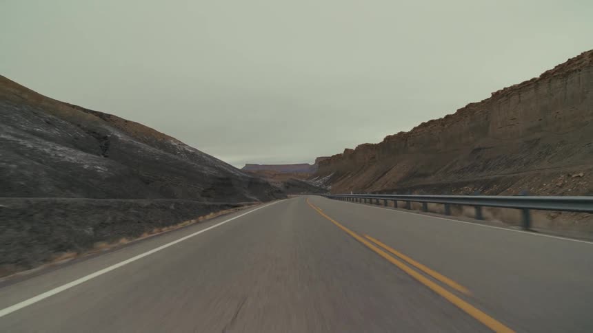 A paved highway winding through a desolate valley of dark rocky mountains and hills under a thick overcast sky