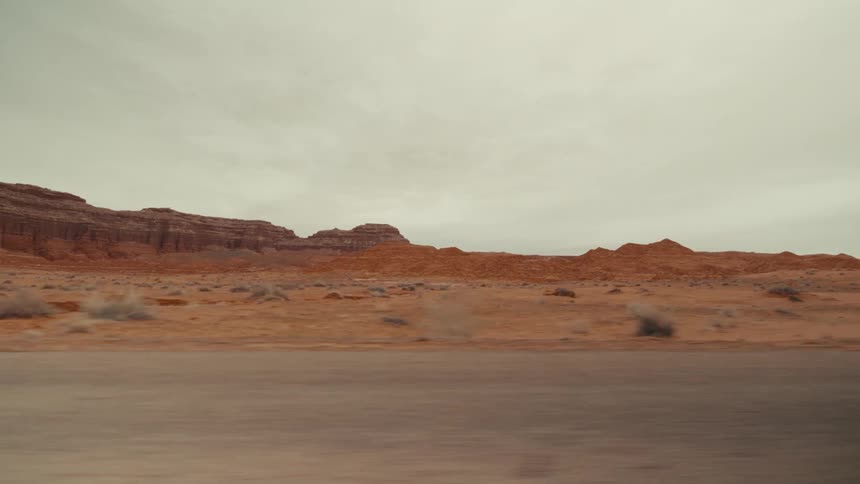 A view from a vehicle moves along a paved road past red rock formations and desert shrubs under a cloudy gray sky