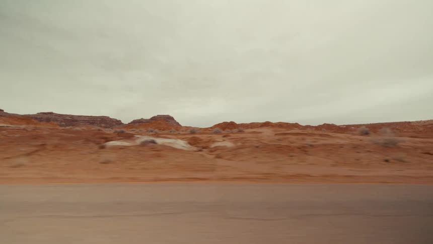 A view from a vehicle moves along a paved road past red rock formations and desert shrubs under a cloudy gray sky
