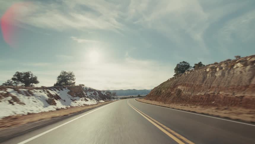 A view from a vehicle moves along a straight paved highway through a dry landscape with scattered green trees and patches of white snow under a blue sky with soft white clouds