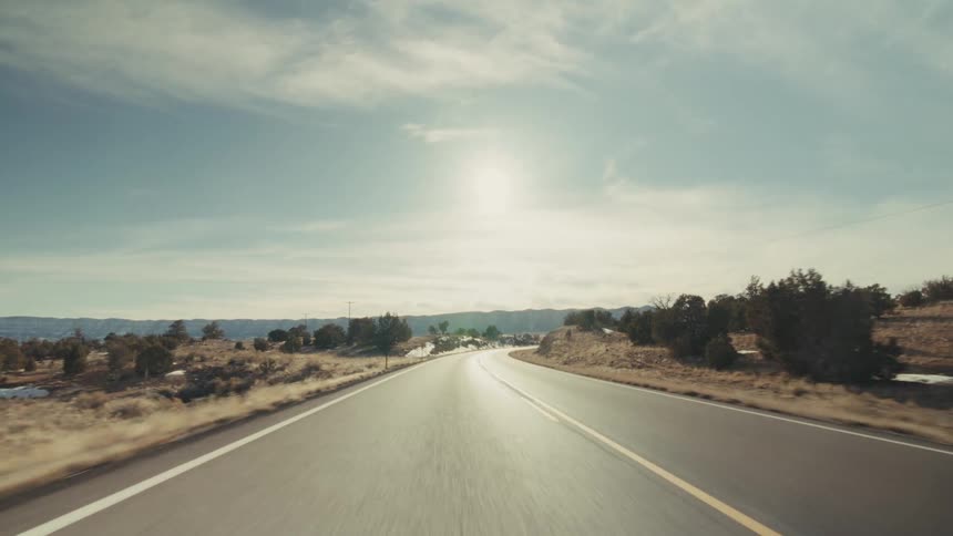 A view from a vehicle moves along a straight paved highway through a dry landscape with scattered green trees and patches of white snow under a blue sky with soft white clouds