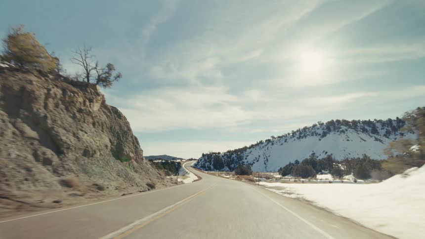 A view from a vehicle travels along a winding two lane road through a mountainous landscape with snowy slopes and green trees under a bright sun and blue sky with wispy clouds