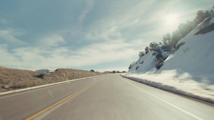A view from a vehicle travels along a winding two lane road through a mountainous landscape with snowy slopes and green trees under a bright sun and blue sky with wispy clouds