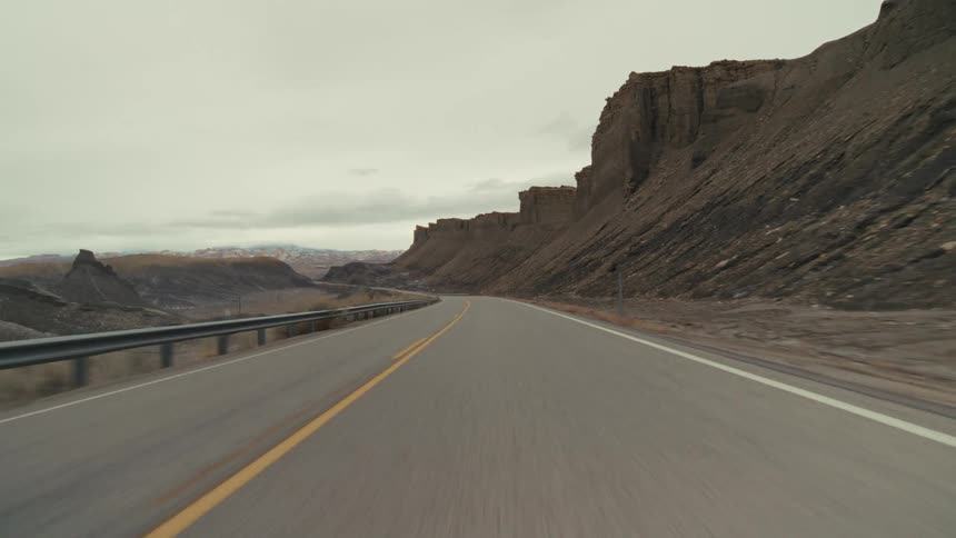 A front facing point of view shot shows a car driving down a two lane road through a mountainous desert landscape under an overcast sky