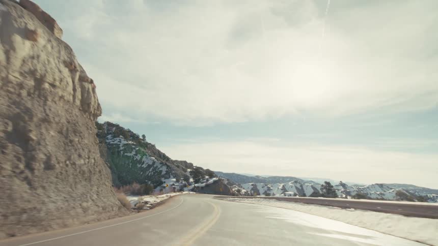 A curved asphalt highway through a vast winter canyon featuring steep rocky hillsides covered in patches of white snow under a bright blue sky with thin clouds and a distant mountain range on the horizon