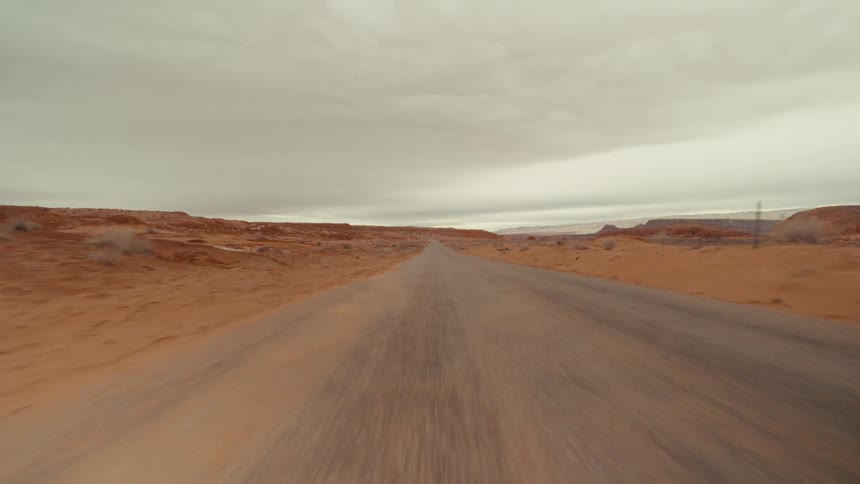 Point of view shot traveling down an asphalt road through a red rock desert landscape under an overcast sky