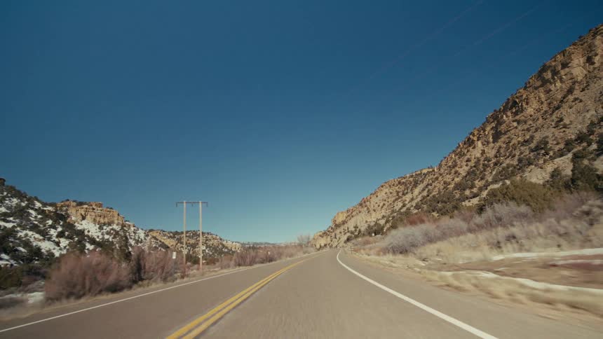 A view from a moving vehicle travels along a curved paved highway with two yellow lines through a rocky canyon with patches of white snow under a blue sky with soft clouds