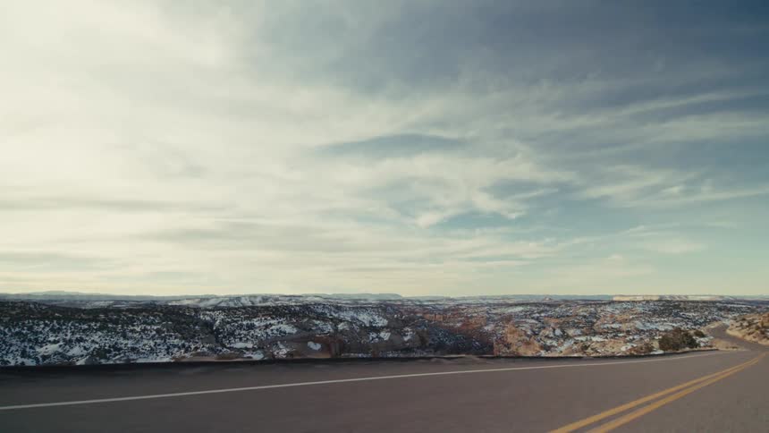 A curved two lane highway that overlooks a vast snow dusted canyon and distant mountain ranges under a bright cloudy sky during a cold winter day in a high desert environment
