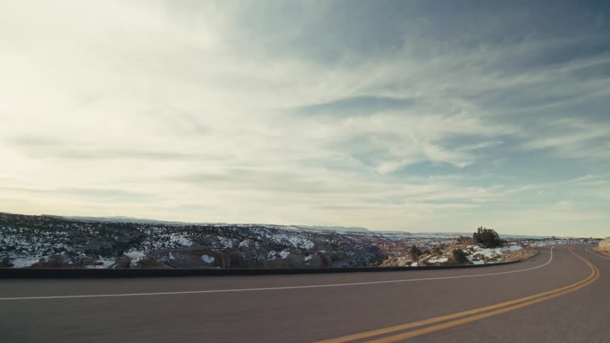 A curved two lane highway that overlooks a vast snow dusted canyon and distant mountain ranges under a bright cloudy sky during a cold winter day in a high desert environment