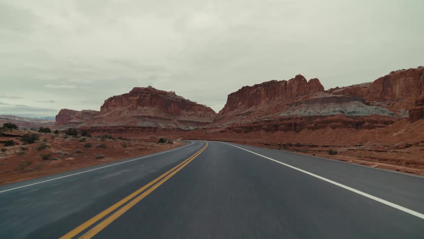 A winding two lane asphalt highway through a vast desert canyon surrounded by massive red rock formations and dry sandy hills under a thick layer of grey clouds
